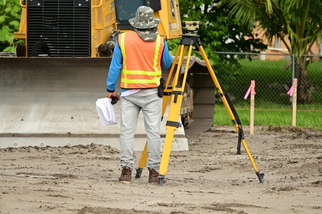 commercial-plumbing Técnico de topografia com colete refletor a trabalhar junto a tripé e nível em estaleiro de construção, realizando medições para mapeamento e levantamento do terreno.